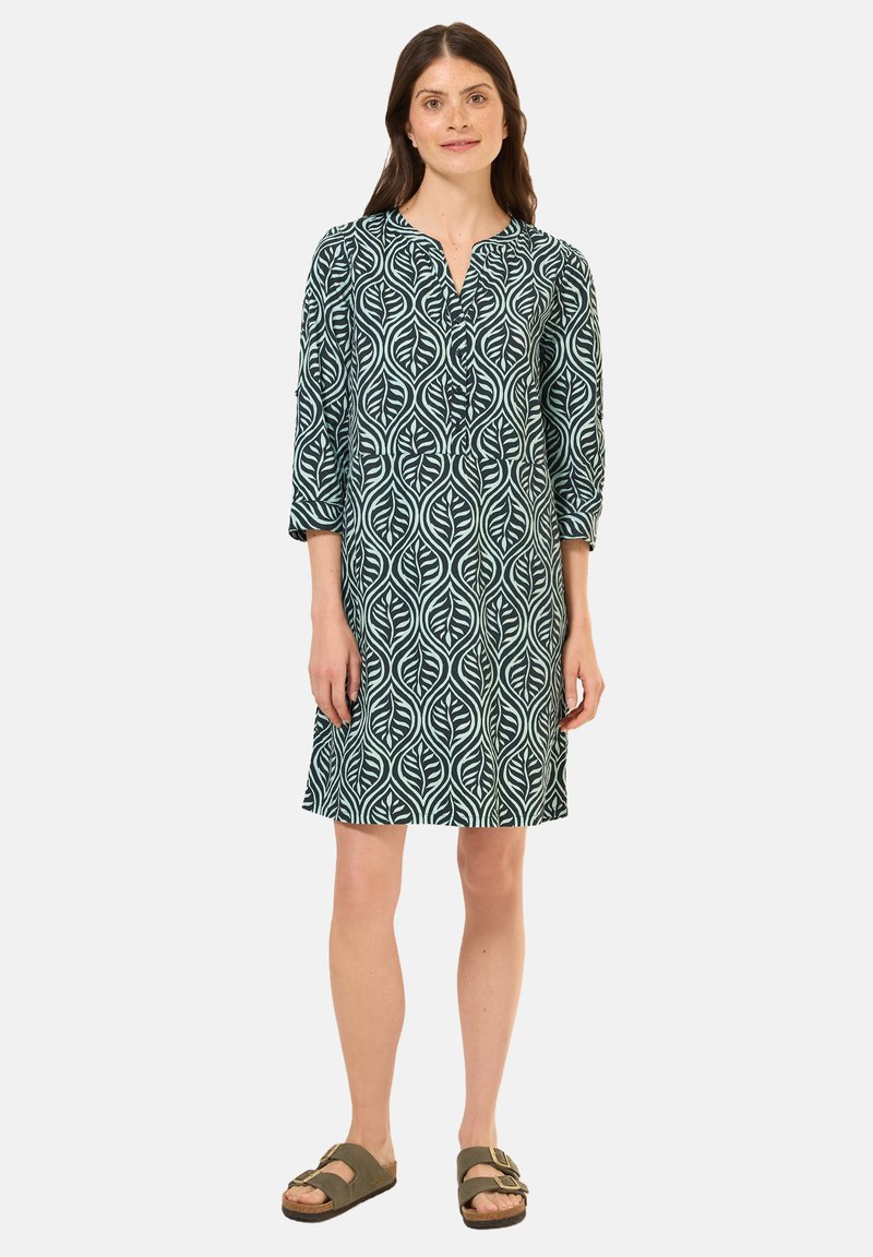 Vihreä mekko, jossa on lehdenkuvio, 3/4-hihat ja kauluskanta. Kangas näyttää kevyeltä ja pehmeältä. Yhdistetty ruskeisiin sandaaleihin.