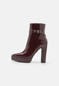 Bottine en cuir verni bordeaux avec un bout pointu, un talon épais et haut, et une sangle décorative avec des clous argentés.