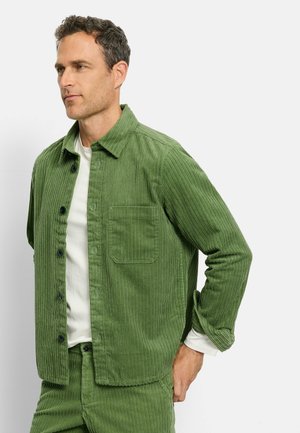 Green corduroy shirt jacket with vertical lines, front button closure, and one chest pocket. Worn over a white shirt.