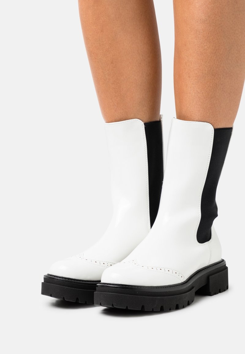 Bottes blanches mi-mollet avec des panneaux latéraux élastiques noirs et des semelles épaisses noires à crampons portées sur des jambes nues sur un fond uni.