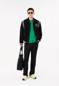Chaqueta varsity negra con rayas blancas, sudadera verde, pantalones negros y zapatillas con tonos amarillos. Llevando una bolsa negra.