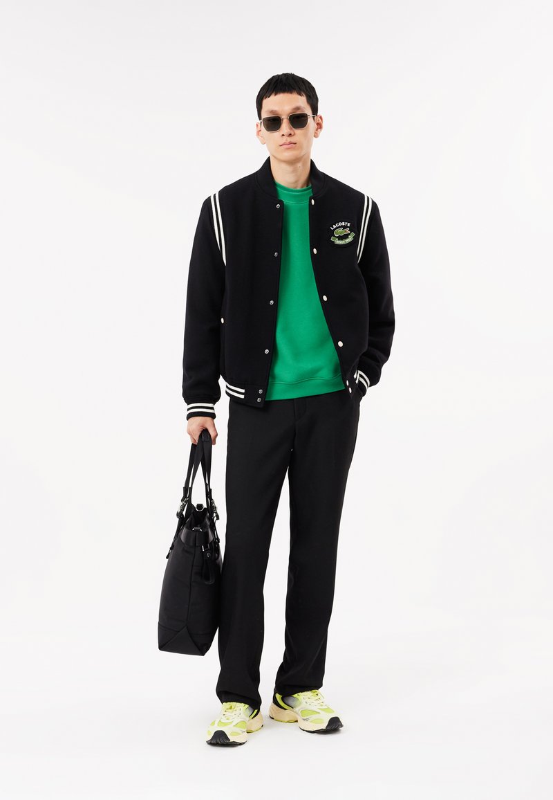 Veste de varsity noire avec des rayures blanches, sweat-shirt vert, pantalon noir et baskets à accents jaunes. Porte un sac noir.