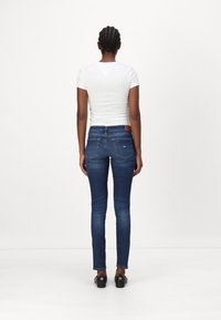 Tmavě modré džíny slim-fit se středně vysokým pasem, zadními kapsami a koženou nášivkou na pásku. Kombinovány s přiléhavým bílým tričkem.