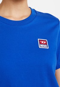 Kungsblå bomullströja med rund halsringning, prydd med en liten fyrkantig röd och vit logotyp som står "DIESEL."