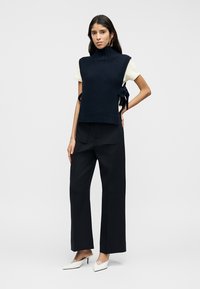 Μπλε σκούρο γιλέκο χωρίς μανίκια με ψηλό γιακά, συνδυασμένο με λευκό T-shirt. Μαύροι παντελόνες με φαρδιά μπατζάκια και λευκά μυτερά παπούτσια ολοκληρώνουν την εμφάνιση.