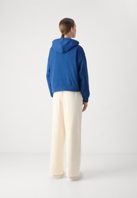 Felpa blu con vestibilità comoda, dotata di cappuccio con cordino. Abbinata a pantaloni larghi crema, calzature bianche e una texture liscia.