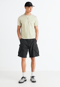 Lichte olijfgroene T-shirt met korte mouwen, gecombineerd met zwarte cargoshorts. Het model draagt een zwarte pet en grijze schoenen en staat tegen een witte achtergrond.