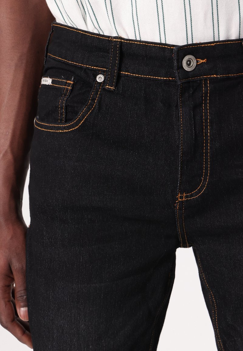 Nahaufnahme einer Person, die schwarze Jeans mit orangefarbenen Nähten und ein weißes Hemd mit grünen Streifen trägt, das in die Hose gesteckt ist.