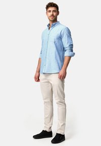 Lys blå skjorte med knapper og brystlomme, parret med beige slim-fit bukser og sorte sneakers, båret af en stående mandlig model.