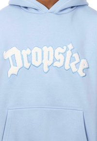 Helle blaue Kapuzenjacke mit einer Fronttasche, die in einer stilisierten Schriftart bold in Weiß und Blau bestickten Text "Dropske" zeigt. Weicher Stoff.