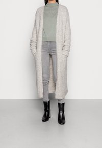 Długi, jasnoszary kardigan o puszystej fakturze, zestawiony z dopasowanym, jasnooliwkowym swetrem i jasnoszarymi rurkami. Całość dopełniają czarne botki za kostkę.