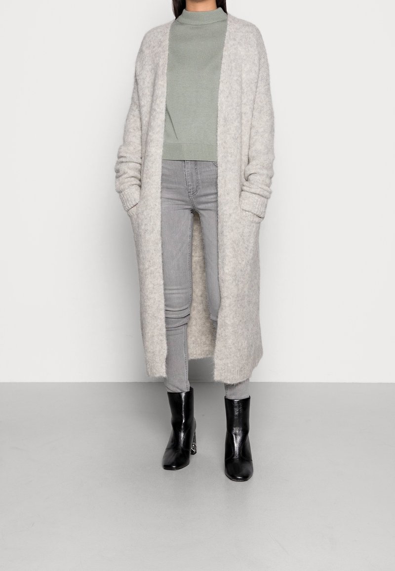 Długi, jasnoszary kardigan o puszystej fakturze, zestawiony z dopasowanym, jasnooliwkowym swetrem i jasnoszarymi rurkami. Całość dopełniają czarne botki za kostkę.