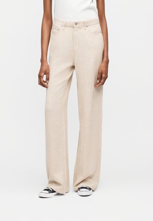 Άτομο που φοράει μπεζ παντελόνι με φαρδιά μπάλα και ασπρόμαυρα αθλητικά παπούτσια, στέκεται μπροστά σε λευκό, απλό φόντο.