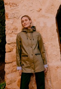 Kobieta o krótkich blond włosach, ubrana w oliwkowo-zielony, kapturowy płaszcz przeciwdeszczowy z zamkiem błyskawicznym i zatrzaskami, stoi na tle teksturowanej kamiennej ściany.