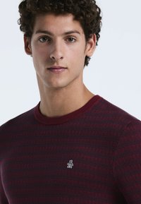 Bordeaux gestrickter Pullover mit Rundhalsausschnitt, der ein dezentes texturiertes Muster und ein kleines Logo auf der Brust aufweist.