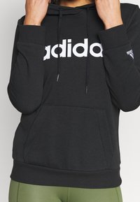 Černá mikina s kapucí vyrobená z měkké látky, která má velké bílé logo "adidas". Obsahuje přední kapsu a šňůrky na kapuci.