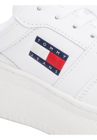 Białe skórzane sneakersy z wyraźnym logo "TOMMY JEANS" w kolorze granatowym i czerwonym, w kształcie prostokąta, oraz teksturowaną gumową podeszwą.