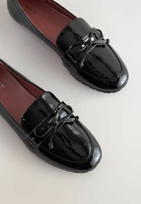 Mocassini in pelle verniciata nera con punta arrotondata, dettaglio decorativo a fiocco e interno bordeaux. Presentano accenti testurizzati e una suola flessibile.