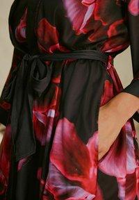 Musta mekko, jossa on kukkakuosi punaisen ja vaaleanpunaisen sävyissä. Varustettu solmittavalla vyöllä ja sivutaskuilla. Silkkinen, kevyt kangas.