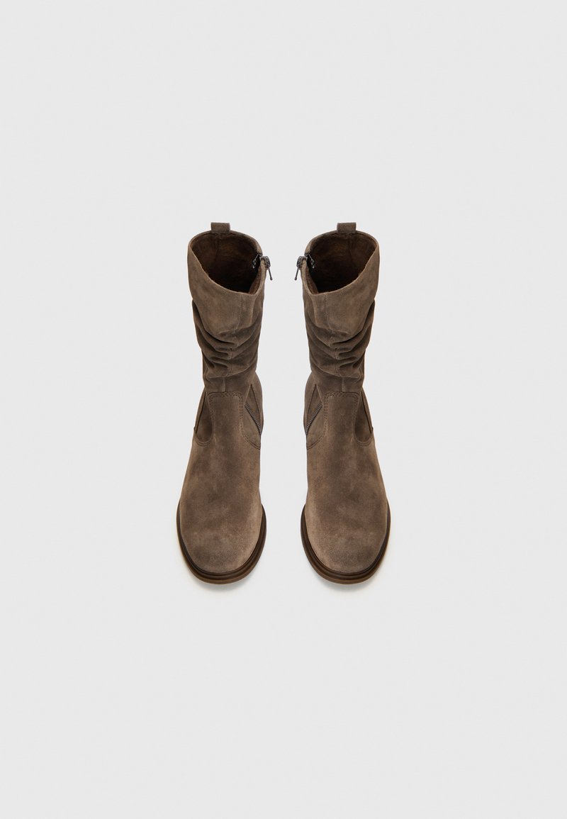 Botines de ante marrón con diseño arrugado, cierre de cremallera lateral y suela ligeramente elevada. Textura suave y puntera redondeada.