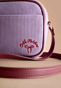 Fialová bavlněná taška s tmavě červeným koženým popruhem a vyšívaným červeným textem. Má lemované okraje a hladkou texturu.