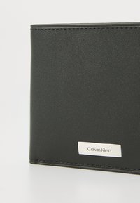 Portefeuille en cuir noir avec une texture lisse, doté d'une plaque logo en argent embossée avec "Calvin Klein" et des bords cousus.