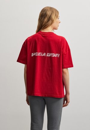 Femme aux longs cheveux blonds portant un t-shirt rouge oversize avec le texte « PEGADOR » au dos, associée à un pantalon gris, vue de dos.