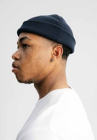 Marineblauwe gebreide beanie met ribbeltextuur, gedragen op het hoofd van een persoon. Witte shirt zichtbaar bij de halslijn. Subtle huidflekken.