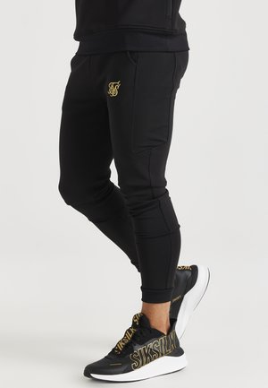 Persoon draagt zwarte taps toelopende joggerbroek met goudkleurig logo en zwarte sneakers met grote gouden merktekens op een witte zool.