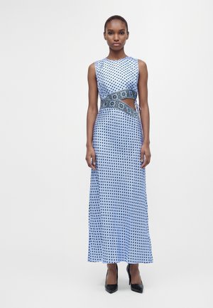 Femme portant une robe maxi bleue sans manches avec de petits motifs carrés noirs et une découpe à la taille, associée à des talons hauts noirs.