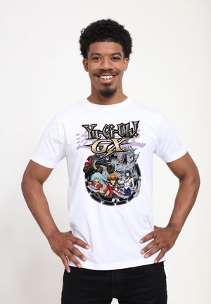 Lachende man die een wit Yu-Gi-Oh! GX-shirt draagt met animefiguren en een mechanisch draakontwerp, staand met de handen in de zij.