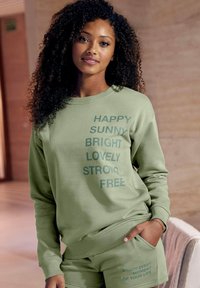 Sudadera verde claro con un diseño de texto en negrita, mangas largas, puños de canalé, y pantalones cortos a juego que cuentan con un bolsillo y un detalle de texto adicional.