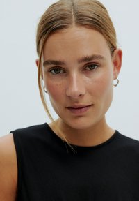 Schwarzes ärmelloses Oberteil mit rundem Ausschnitt, glatter Textur und minimalistischem Design, getragen von einer Person mit braunen Haaren und kreisförmigen Ohrringen.