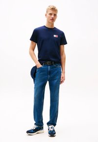 Jaunas vyras, dėvintis tamsiai mėlyną marškinėlį su logotipu, mėlynas džinsus ir tamsiai mėlynas sportbačius, stovintis viena ranka kišenėje ant baltos fono.