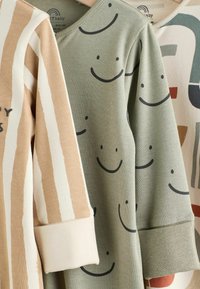Trois vêtements pour bébé à manches longues avec des poignets roulés, un beige rayé blanc, un vert avec des visages souriants noirs, et un avec des formes abstraites.