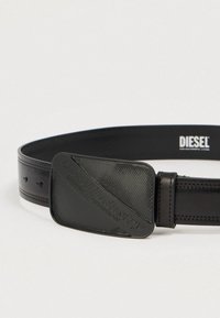 Μαύρη δερμάτινη ζώνη με διακοσμητική αγκράφα που φέρει το ανάγλυφο κείμενο "Diesel Industry Division." Διαθέτει τέσσερις ρυθμιζόμενες τρύπες.