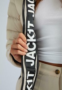 Beige Daunenjacke mit schwarzem Reißverschluss, auf dem große weiße Schriftzüge mit "JACKIT" zu sehen sind. Nahaufnahme der Stoffstruktur und einer Hand, die den Reißverschluss greift.