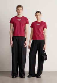 Vínové tričko s krátkým rukávem s nápisem "TOMMY" na hrudi, doplněné černými širokými kalhotami. Žena drží malou černou tašku.