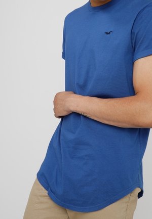 Niebieski bawełniany t-shirt z okrągłym dekoltem i krótkimi rękawami, z małym czarnym logo na lewej piersi, o dopasowanym kroju i zaokrąglonym dolnym brzegu.