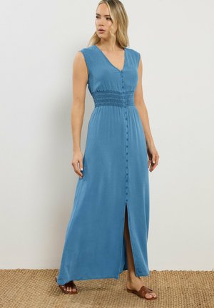 Kvinde i ærmeløs blå maxikjole med knapper foran og frontslids, stående på flettet måtte mod en ensfarvet hvid væg.