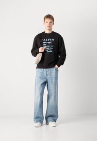 Sweatshirt preto com texto gráfico azul, jeans de perna larga azul claro e tênis brancos. Modelo segura uma bolsa crossbody bege.