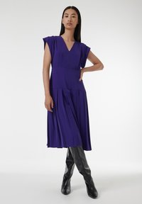 Paarse geplooide jurk met een V-hals, korte mouwen en een getailleerde taille, gedragen met hoge, zwarte, puntige laarzen. Gladde stofstructuur.
