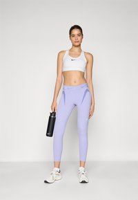Lyse lilla tights med mesh-paneler, hvid sportsbh med sort logo og sort vandflaske, parret med hvide træningsskov. 