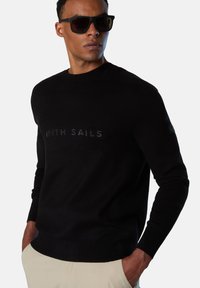 Suéter negro de punto con cuello redondo y logo de "NORTH SAILS" en impresión oscura. Combinado con pantalones beige.