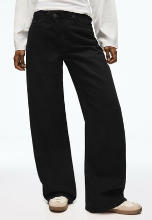 Personne portant un jean noir ample, une chemise blanche à manches longues et des baskets blanches avec des accents beiges.