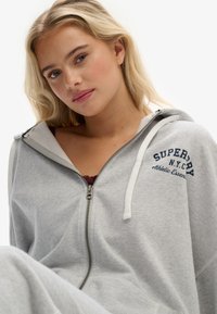 Lys grå zip-up hoodie lavet af blødt stof, med en hvid snor, et logo i marineblå og en afslappet pasform.