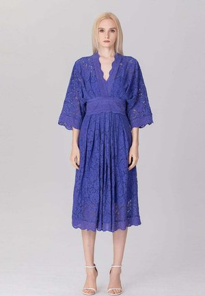 Femme debout portant une robe en dentelle bleue longueur genou avec des manches larges et une taille cintrée, assortie à des talons blancs à bride à la cheville.