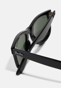 Gafas de sol Ray-Ban negras con montura angular, lentes verde oscuro y un acento de logo plateado en las patillas. Textura suave.