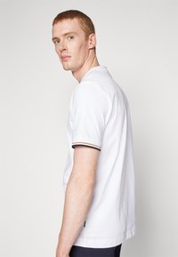 Weißes Baumwoll-T-Shirt mit kurzen Ärmeln, geripptem Kragen mit schwarzen und goldenen Streifen, lockerer Schnitt und seitlichem Etikett.