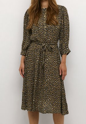 Femme portant une robe midi boutonnée à manches longues à imprimé léopard, nouée à la taille, sur un fond uni.